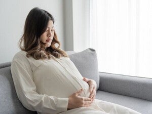 Mulher grávida pensativa sentada no sofá com as mãos na barriga.