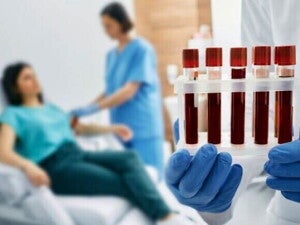 Close-up de uma mão enluvada segurando uma grade com cinco tubos de ensaio contendo sangue. Ao fundo, em desfoque, uma profissional de saúde faz uma coleta de sangue no braço de uma mulher sentada em uma cadeira de hospital.