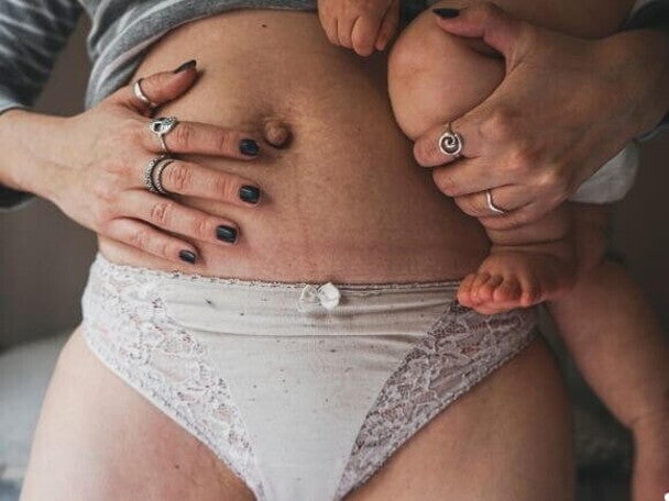 Close na barriga de uma mulher no pós parto usando uma calcinha de cor clara, segurando seu bebê no colo e apoiando outra mão na barriga.