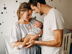 Um homem e uma mulher sorridentes seguram um bebê nos braços, dentro de um quarto infantil.