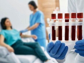 Close-up de uma mão enluvada segurando uma grade com cinco tubos de ensaio contendo sangue. Ao fundo, em desfoque, uma profissional de saúde faz uma coleta de sangue no braço de uma mulher sentada em uma cadeira de hospital.