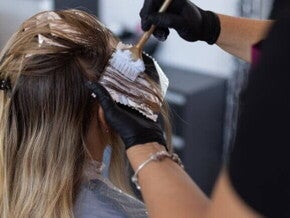 Mãos com luvas aplicando tinta no cabelo loiro de uma mulher, usando pincel e papel alumínio para mechas.