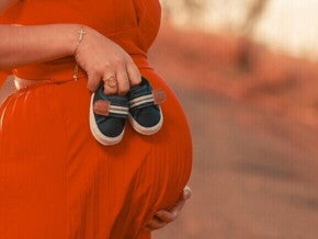 Close-up da barriga de uma mulher grávida, segurando sapatinhos de bebê, simbolizando a espera pelo nascimento.