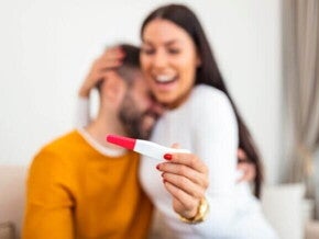 Mulher feliz segurando um teste de gravidez na mão e sendo abraçada por um homem, sugerindo comemoração pelo resultado positivo