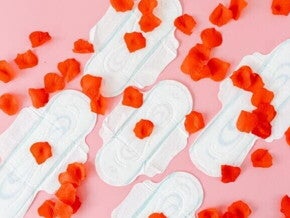 Quatro absorventes higiênicos dispostos sobre um fundo rosa, com pétalas de rosa vermelhas espalhadas.