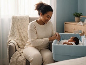 Mãe sentada no sofá enquanto olha sorrindo para o bebê deitado no moisés azul em um quarto de tons claros.