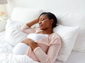 Mulher grávida deitada na cama, de lado, com os olhos fechados. A mão dela está na testa. A imagem mostra a posição para gestantes dormir, com a mulher buscando o conforto necessário para o sono. Mulher grávida deitada na cama, de lado, com os olhos fechados. A mão dela está na testa. A imagem mostra a posição para gestantes dormir, com a mulher buscando o conforto necessário para o sono.
