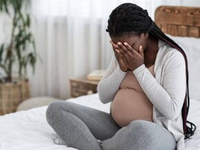 Mulher grávida sentada na cama com as mãos cobrindo o rosto, expressando mal-estar ou dor de cabeça intensa.