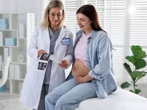 Médica mostrando uma imagem de ultrassom para uma gestante sentada, que toca a barriga, ilustrando um momento chave do acompanhamento na primeira consulta.