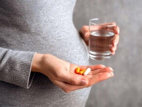 Mulher grávida, de perfil, segura com uma mão um copo de água e, com a outra, um punhado de pílulas coloridas, incluindo cápsulas brancas e amarelas. A imagem destaca a importância de tomar as vitaminas pré-natais para a saúde da mãe e do bebê.