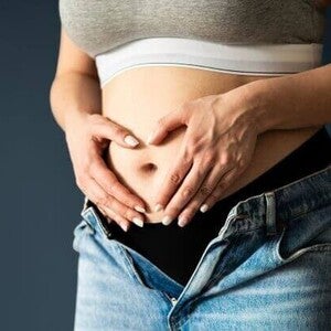 Abdômen de uma mulher grávida no primeiro ou segundo trimestre, mãos em formato de coração sobre a barriga.