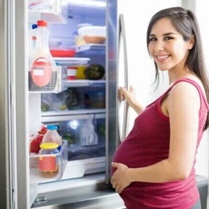 Mulher grávida, segurando a barriga, abre a porta da geladeira em sua cozinha e sorri para a câmera. A imagem ilustra a atenção a ser dada à dieta e aos alimentos para não comer na gravidez.