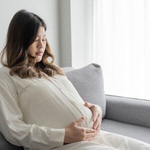 Mulher grávida pensativa sentada no sofá com as mãos na barriga.