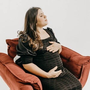 Mulher grávida em um vestido preto sentada em uma poltrona vermelha, segurando a barriga com os olhos fechados.