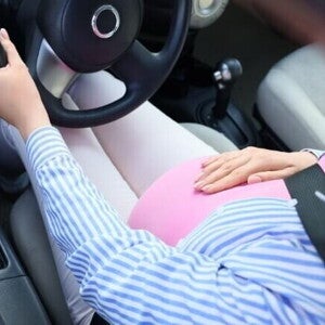 Visão de cima de uma mulher grávida dirigindo um carro, segurando o volante com uma mão e a outra sobre a barriga saliente. O cinto de segurança está posicionado.