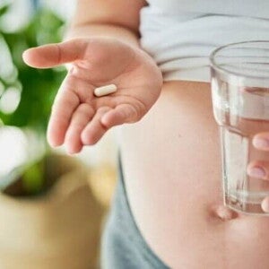Mulher grávida, de perfil, segura uma cápsula em uma mão e um copo de água na outra, para tomar um suplemento. A imagem representa o uso de colágeno para gestante.