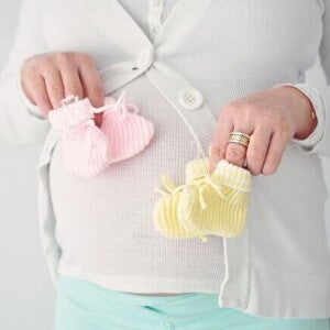 Uma mulher grávida segura dois pares de sapatinhos de bebê, um rosa e um amarelo. A imagem simboliza a gravidez de gêmeos.