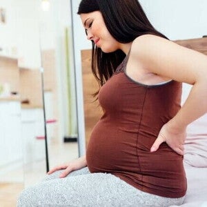Mulher grávida sentada na beira da cama, com expressão de desconforto e a mão apoiada na região lombar.
