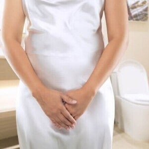 mulher em pé, com as mãos cobrindo a região pélvica, vestindo camisola branca, em banheiro com vaso sanitário ao fundo.