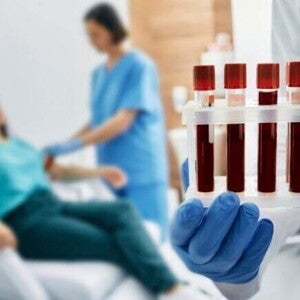 Close-up de uma mão enluvada segurando uma grade com cinco tubos de ensaio contendo sangue. Ao fundo, em desfoque, uma profissional de saúde faz uma coleta de sangue no braço de uma mulher sentada em uma cadeira de hospital.