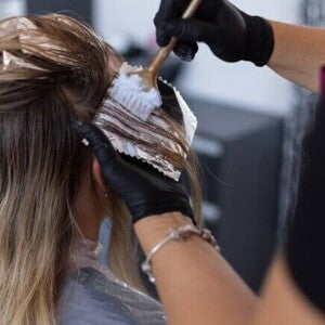 Mãos com luvas aplicando tinta no cabelo loiro de uma mulher, usando pincel e papel alumínio para mechas.