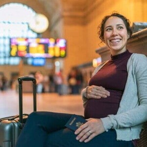 Foto de uma gestante sorrindo e relaxada, sentada em um banco de estação, com o passaporte na mão, indicando viagem internacional.