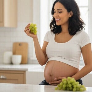 Mulher grávida com a mão na barriga segurando um cacho de uvas sentada em uma cozinha de tons claros