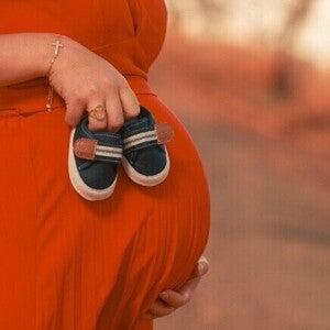 Close-up da barriga de uma mulher grávida, segurando sapatinhos de bebê, simbolizando a espera pelo nascimento.