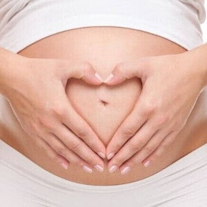 Close de mulher grávida com as mãos formando um coração sobre a barriga.