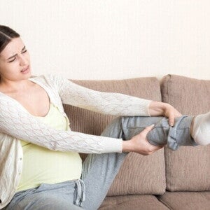 Gestante sentada no sofá, com expressão de dor, massageando a perna devido ao inchaço na gravidez comum nos membros inferiores.