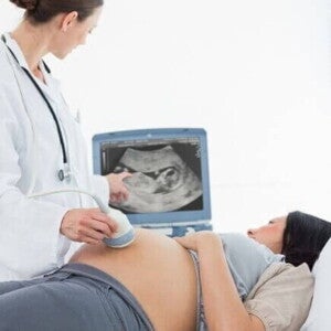 Médica realiza exame de ultrassonografia em mulher grávida para diagnóstico de líquido amniótico aumentado. A imagem mostra o monitor do aparelho exibindo o feto e a médica apontando para a tela.