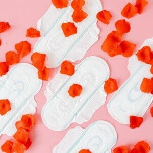 Quatro absorventes higiênicos dispostos sobre um fundo rosa, com pétalas de rosa vermelhas espalhadas.