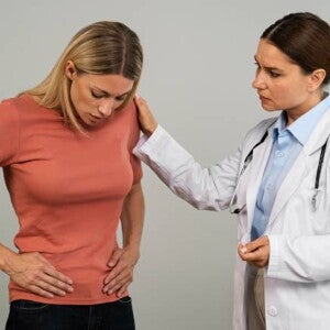 mulher vestindo camiseta laranja com as mãos posicionadas na barriga, ao lado direito uma mulher vestindo jaleco branco mulher vestindo camiseta laranja com as mãos posicionadas na barriga, ao lado direito uma mulher vestindo jaleco branco