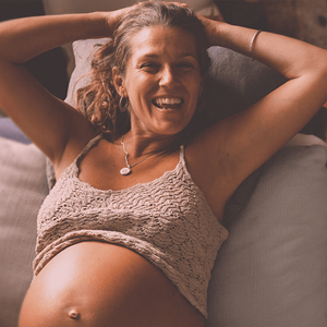 Pregnant woman smiling, with her arms behind her head and top raise to reveal her belly.