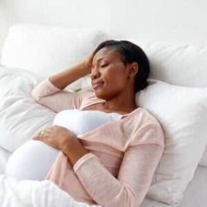 Mulher grávida deitada na cama, de lado, com os olhos fechados. A mão dela está na testa. A imagem mostra a posição para gestantes dormir, com a mulher buscando o conforto necessário para o sono.