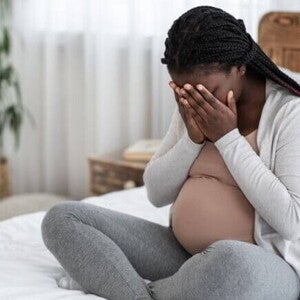 Mulher grávida sentada na cama com as mãos cobrindo o rosto, expressando mal-estar ou dor de cabeça intensa.