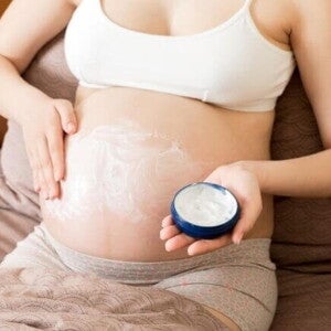 Mulher grávida sentada na cama aplicando um creme hidratante branco na barriga para aliviar o prurido na gravidez. Ela segura um pote azul de creme em uma das mãos enquanto espalha o produto na pele esticada com a outra