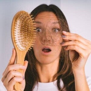 Uma mulher com expressão de surpresa segura um tufo de cabelo que se desprendeu na escova. A imagem representa o choque e a preocupação com a queda de cabelo pós-parto.
