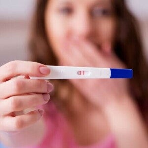 Mulher com expressão de surpresa, segurando um teste de gravidez que mostra o resultado positivo.