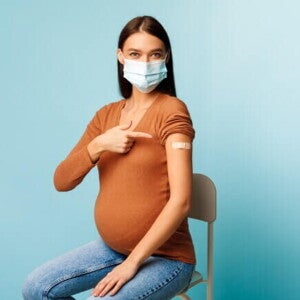 Mulher grávida com máscara apontando para o curativo no braço após receber uma das vacinas e gravidez recomendadas, simbolizando a proteção durante a gestação.