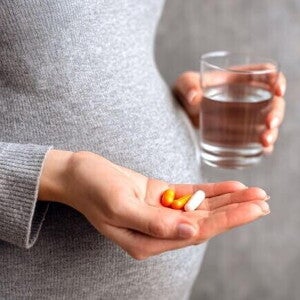 Mulher grávida, segurando na mão um copo de água e três cápsulas/pílulas, exemplificando a suplementação de vitaminas para gestante.