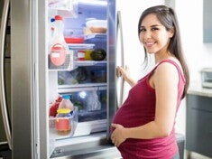 Mulher grávida, segurando a barriga, abre a porta da geladeira em sua cozinha e sorri para a câmera. A imagem ilustra a atenção a ser dada à dieta e aos alimentos para não comer na gravidez.
