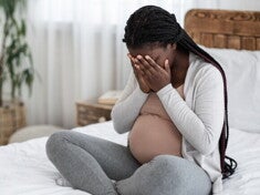 Mulher grávida sentada na cama cobrindo o rosto com as mãos.