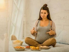 grávida aponta para a barriga sentada em uma cama, rodeada de pratos variados.