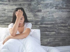 mulher sentada em cama com lençóis brancos enquanto está cabisbaixa.