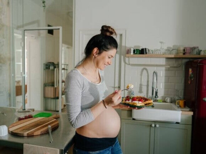 Mulher grávida sorrindo na cozinha enquanto come um doce