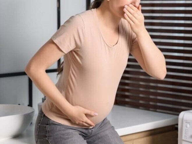 Mulher grávida em pé, vestindo camiseta bege, com a mão na boca e a outra no abdômen, expressando desconforto e ânsia na gravidez.