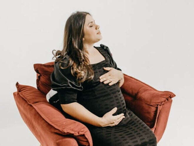 Mulher grávida em um vestido preto sentada em uma poltrona vermelha, segurando a barriga com os olhos fechados.