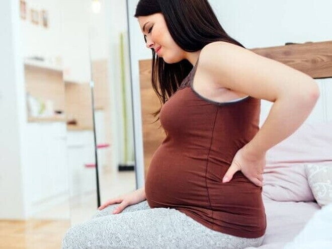 Mulher grávida sentada na beira da cama, com expressão de desconforto e a mão apoiada na região lombar.