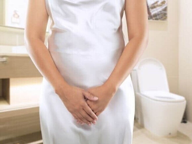 mulher em pé, com as mãos cobrindo a região pélvica, vestindo camisola branca, em banheiro com vaso sanitário ao fundo.